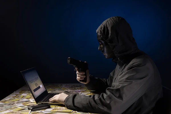 Anonymous hacker programmer uses a laptop holding a gun in his hand to hack the system in the dark. Creation and infection of malicious virus. — Stock Photo, Image