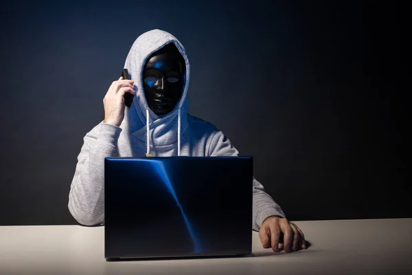 Anonyme Hacker in Maske Programmierer verwendet einen Laptop und telefoniert, um das System im Dunkeln zu hacken. — Stockfoto