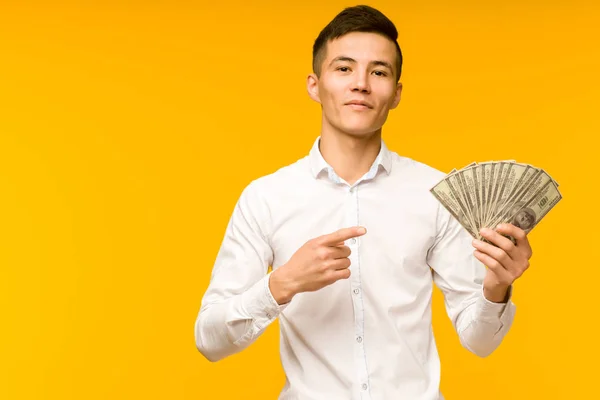 Joyful Asian man i en vit skjorta pekar ett finger på pengar dollar på en gul bakgrund — Stockfoto