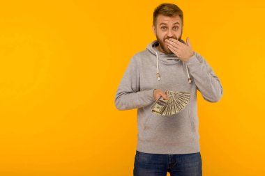 Gri kapüşonlu neşeli adam sarı bir arka plan üzerinde eliyle ağzını kaplayan para dolar tutar