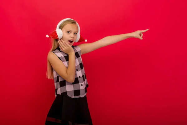 Cheerful little girl pointing her fingers . A child in a warm ear muffs on a red background.