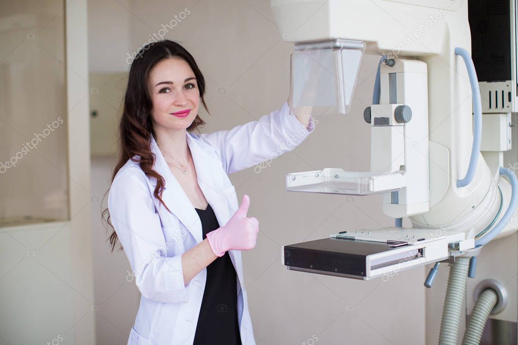 Portrait of the young smiling breast specialist standing near apparatus of the ultrasound examination of the breast in her office and showing thumb up