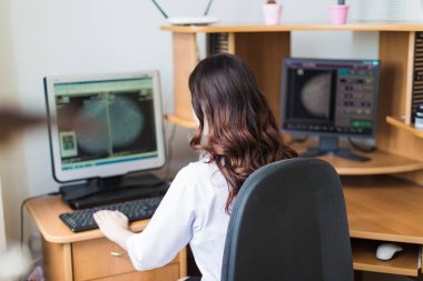 Back view of the young breast specialist who is sitting at the computer and looking at the results of the ultrasound examination in her office clipart