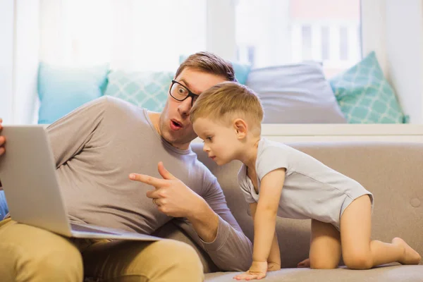 The surprised young father sitting on a sofa in the room near his cute little son and they watching laptop