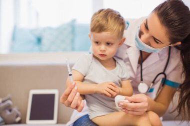 Genç hemşire odasında kanepede oturan ve sevimli küçük çocuk üstünde belgili tanımlık kucak tutarak ve şırınga elinde tutan