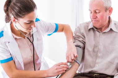 Bir yatak odasında evde ve genç ciddi hemşire yanına oturmuş ve kan basıncı ölçüm üzerinde oturan yaşlı hasta adam