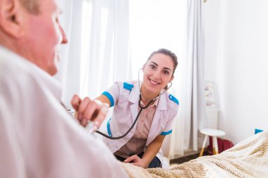 Yaşlı hasta adam evde bir yatakta yatarken ve bir steteskop kullanarak gürültü dinleme yakın oturan genç gülümseyen doktor