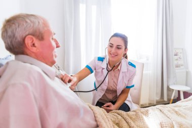 Yaşlı hasta adam evde bir yatakta yatarken ve bir steteskop kullanarak onun organizma gürültü dinlerken yakın oturan genç gülümseyen doktor