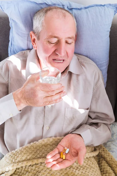 Portrait of the old sick man lying on a bed and holding pills and glass of water in hands at home