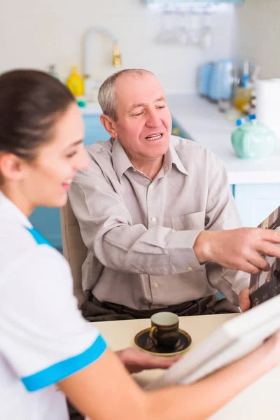 病気の老人と自宅のフォト アルバムを確認彼らの近くのテーブルに座っている若い笑顔の医者 — ストック写真