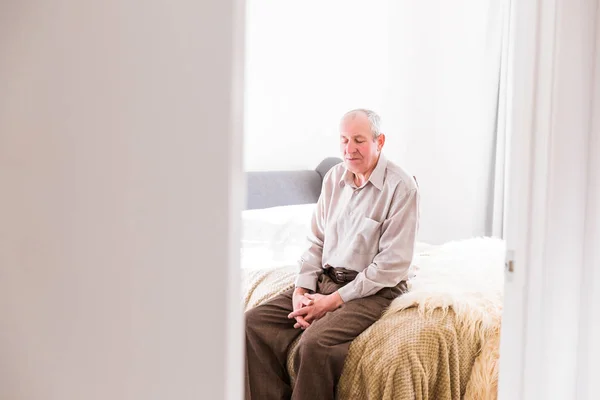 The old man sitting on a bed in the room at home and looking down
