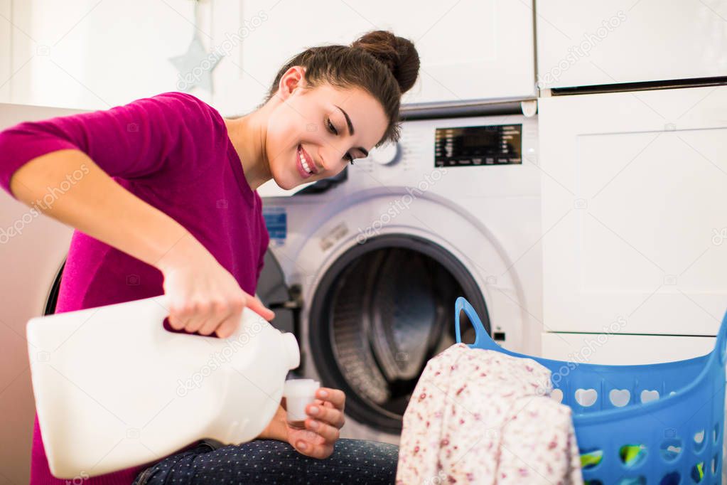 Portrait of the young smiling woman who sitting near washing machine in the room and pouring rinses into the lid