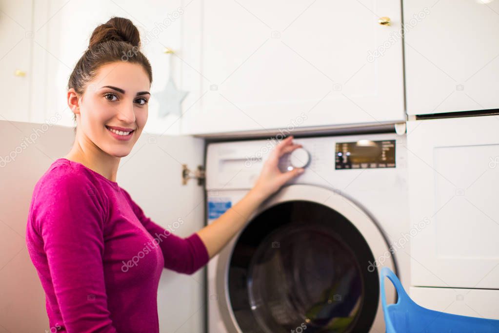 The young smiling woman sitting near washing machine in the room and turning on the washing machine