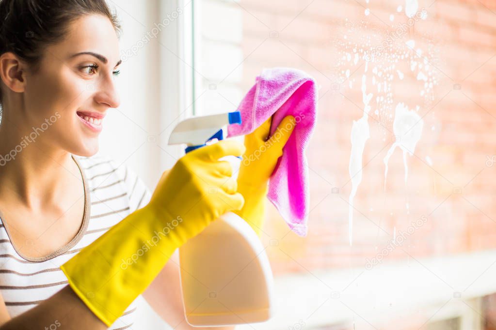 The young cheerful woman in gloves washing the window at home