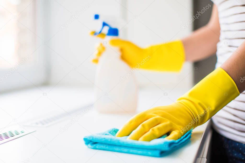 Close-up the young woman in yellow gloves holding rag and window cleaner in hands near the window indoors