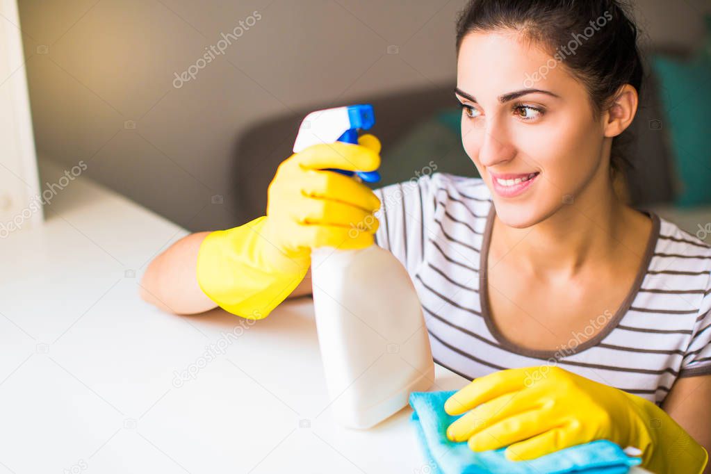 Portrait of the young smiling woman in yellow gloves who holding rag and window cleaner in hands and looking in the window in the room