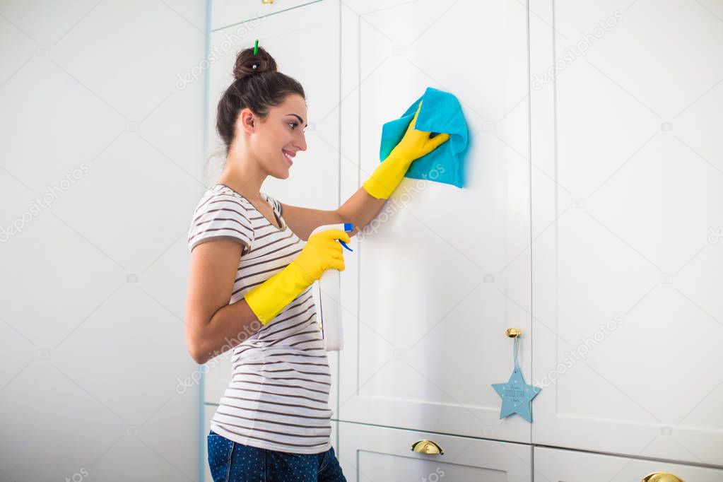 Side view of the young smiling woman in yellow gloves who holding rag and detergent in hands and washing furnitures in the room