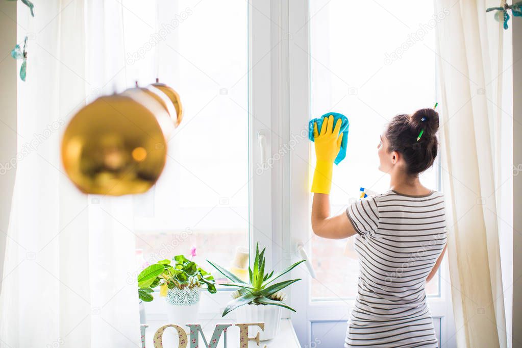 Back view of the young woman in yellow gloves who holding rag and window cleaner in hands and washing the window in the room