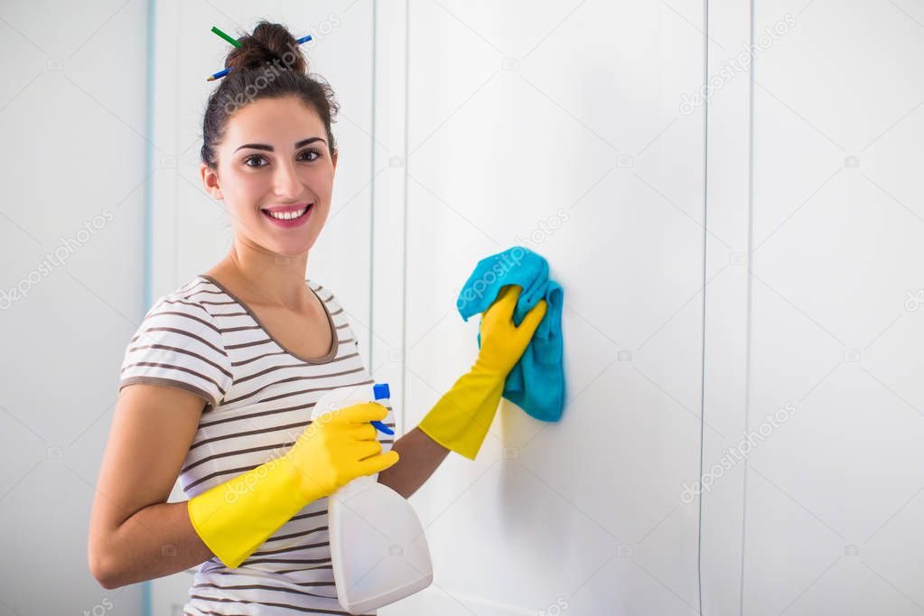 Portrait of the young smiling woman in yellow gloves who holding rag and detergent in hands and washing furnitures in the room