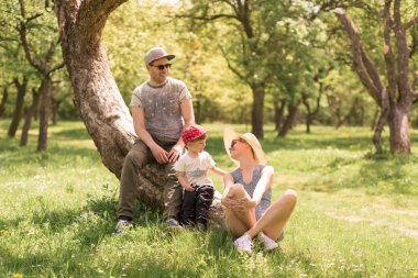 Neşeli küçük çocuk ve mutlu baba bir ağaç ve çim üzerinde oturan anne oturan yakın onları içinde belgili tanımlık bahçe ve gülüyorlar