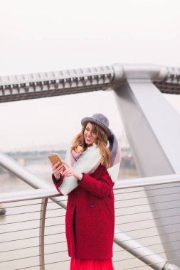 Genç güzel gülümseyen Bayan şapka ayakta açık ve telefon bakarak köprüde