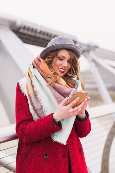 Portrait of young beautiful joyful lady in hat standing on the bridge outdoor and looking at the phone