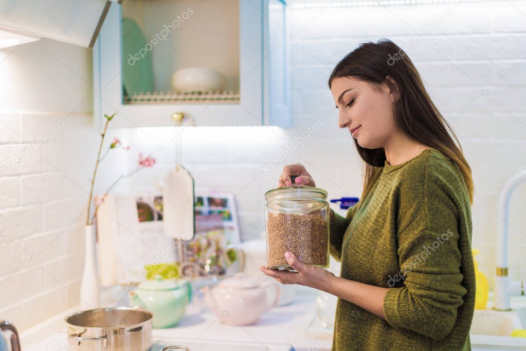 Side view of the young woman who cooking in the kitchen and holding dish with buckwheat in hands