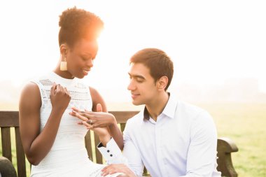 Çift güneş ışığı açık bir bankta oturmuş ve birbirlerine elele gülümseyen genç romantik portresi