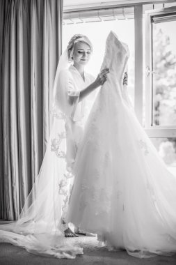 Genç mutlu güzel gelin odanın penceresinde yakın duran ve gelinlik elinde tutan ve bakarak düğün peçe