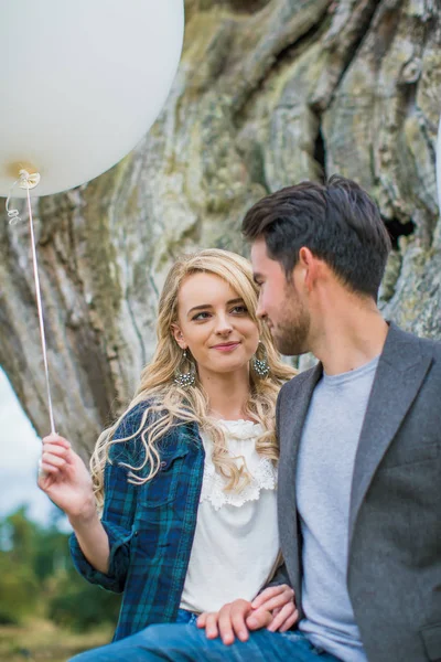 年轻美丽的微笑的夫妇谁站在户外 依靠老树干的树和手持白色气球在手的肖像 — 图库照片