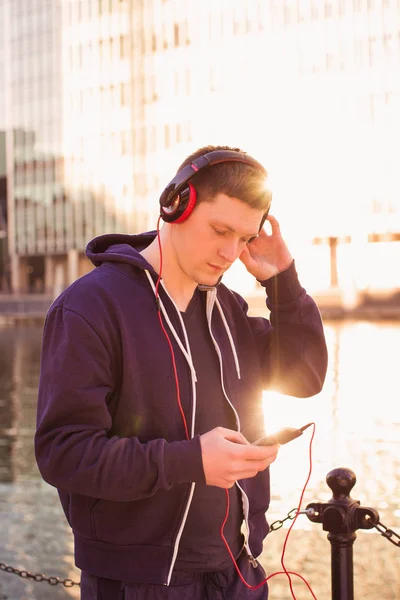 Portrait of young sporty man who standing outdoor and listen to music in the earphones and using the phone