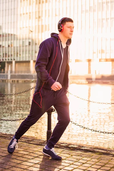 The young cheerful sporty man running in the sunlight by the river and listen to music in the earphones