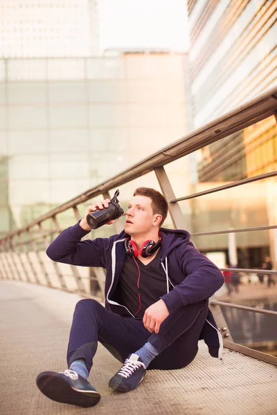 The young sporty man sitting on the floor outdoor and drinking the water