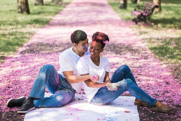 The young smiling couple sitting with paint on their faces on the road in the park and holding the dry paint in hands