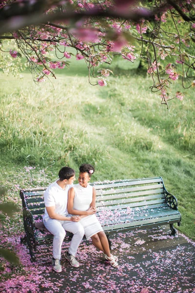 花弁が公園でベンチに抱擁で座っているとお互いを見て 笑みを浮かべて愛の若いカップル — ストック写真