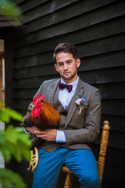 The young happy handsome fiance sitting on the chair near home and holding chicken in hands