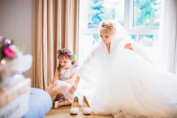 美しい幸せな花嫁の結婚式を保持している部屋で手に服装をするいると窓の近くの床に座って美しい少女 — ストック写真