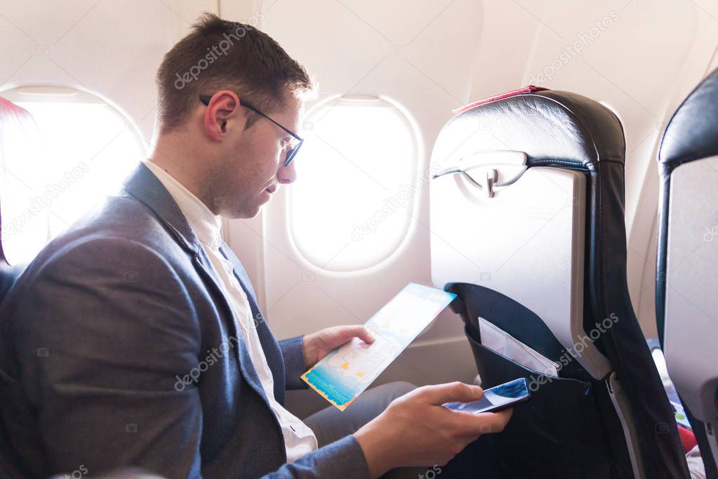 The elegant young man sitting on the plane near the window and reading and holding the phone in hand