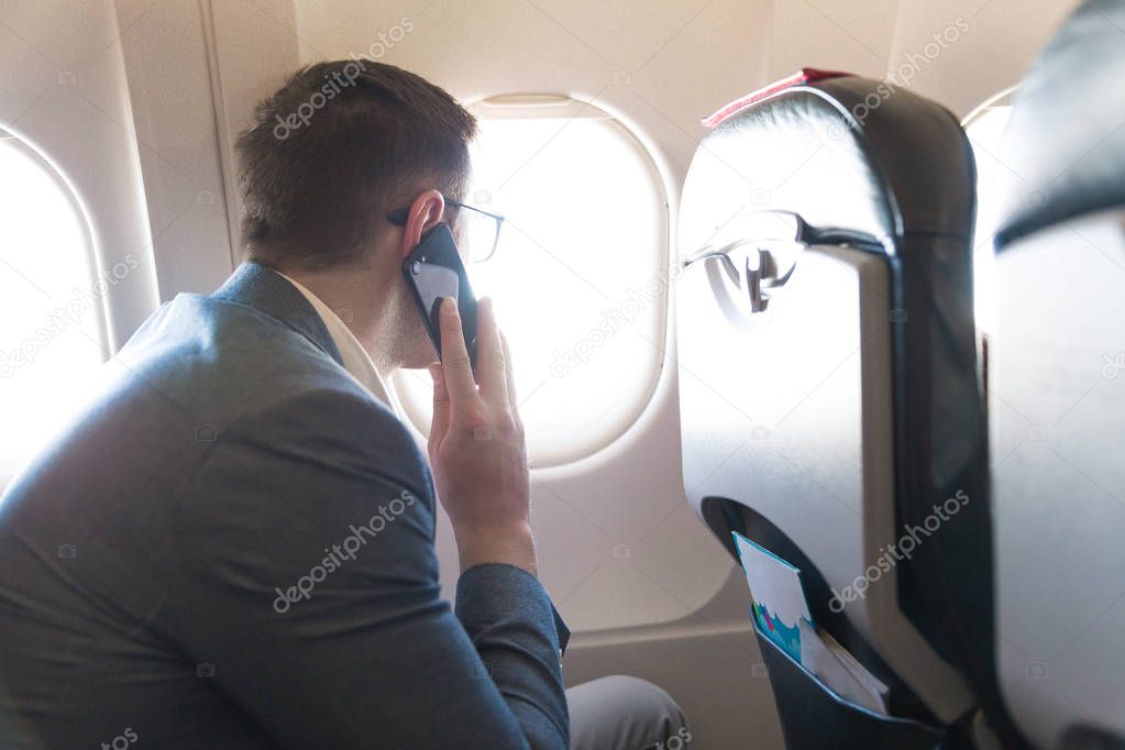 The elegant young man sitting on the plane near the window and using the phone