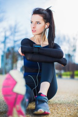 Sportif kadın yolda oturur, bir kenara görünüyor ve parkta kulaklık müzik dinler