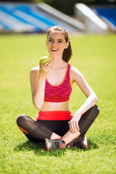运动礼服的运动微笑的妇女坐在体育场和保持一个苹果在手 — 图库照片