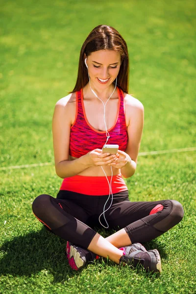 Young sporty joyful woman in sport dress sits on the grass and listens to music in earphones