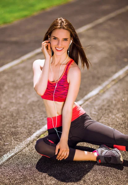 Young sporty smiling woman in sport dress sits on stadium track and listens to music in earphones