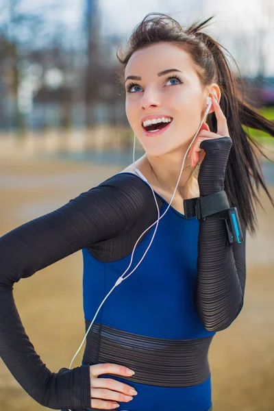 Happy attractive sporty woman stands and listens to music in earphones, in the park