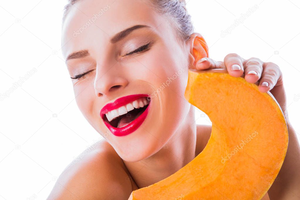 Close-up young smiling woman with bare shoulders and closed eyes keeps pumpkin in hands near her face on the white background