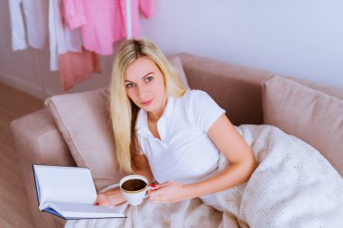 Kanepe tutar fincan kahve ve oda onun elinde bir kitap üzerinde çekici sarışın kadın yatıyor