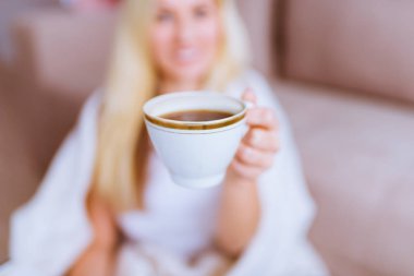Gülümseyen sarışın kadın kanepede oturur ve ön planda onun elinde kahve tutar