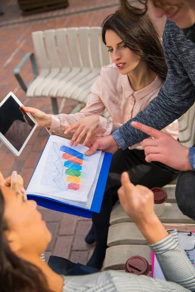 Clever students sit on a bench with the tablet and folder in their hands, discuss their tasks in time of a break, outdoors