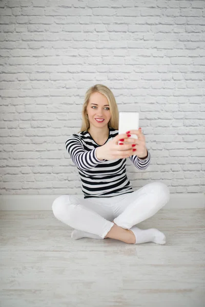 Attractive blonde woman sits on the floor, keeps the phone in her hands and makes selfie-photo, at home