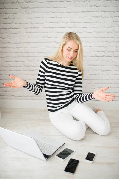 A wonderful blonde woman sits on the floor with three phones and laptop, looks at gadgets and shows of gesture that she does not know what phone she needs, in the room